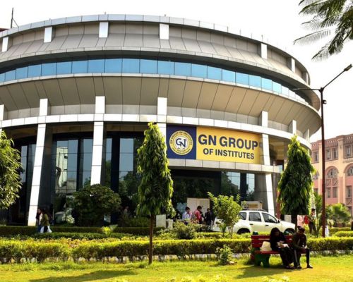 A modern circular academic building of GN Group of Institutes on a sunny day. A yellow sign is above the glass door, with a large park, red benches, and students in the foreground.